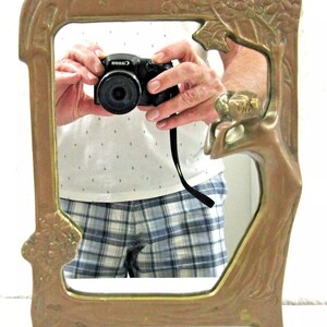 May include: A vintage brass mirror with a decorative frame featuring a stylized tree design with floral accents. The mirror has a rectangular shape and stands on two feet.