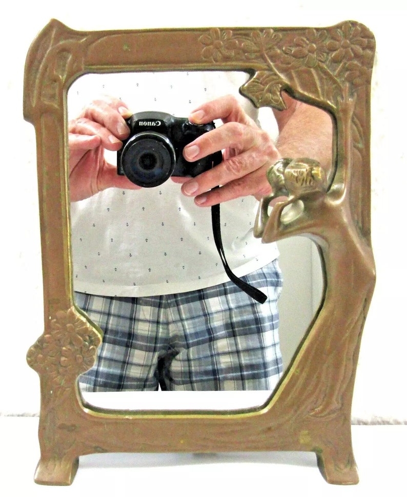 May include: A vintage brass mirror with a decorative frame featuring a stylized tree design with floral accents. The mirror has a rectangular shape and stands on two feet.