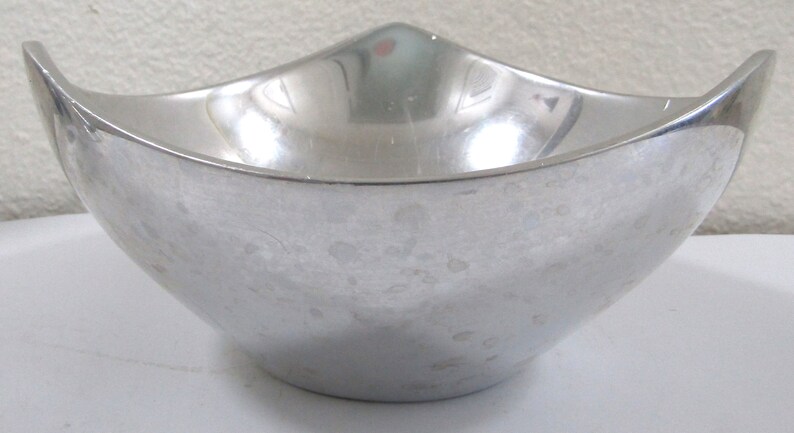 Puede incluir: Un cuenco de metal plateado con un dise&ntilde;o curvo &uacute;nico. El cuenco tiene una superficie lisa y reflectante con motas sutiles.