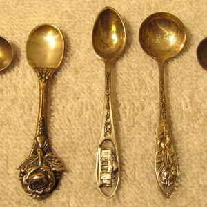 May include: Five silver teaspoons with ornate designs. The spoons have different designs on the handles, including a rose, a building, and a floral pattern.