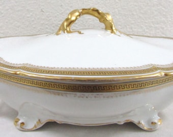 May include: A white porcelain serving dish with a lid, featuring gold trim and handles. The dish has a decorative Greek key pattern around the rim and on the lid. The dish is oval-shaped and rests on three small feet.
