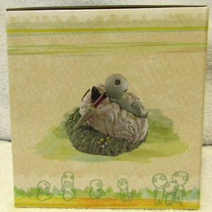 May include: A small, gray, ceramic figurine of a creature with a large head and a small body. The creature is lying on its back on a white, fluffy cloud. The cloud is surrounded by green grass and small, white flowers. The figurine is part of a set of similar figurines.