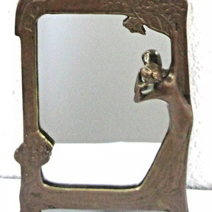 May include: A bronze mirror frame with an Art Nouveau design featuring a woman holding a flower. The frame has a decorative floral pattern and stands on a base.