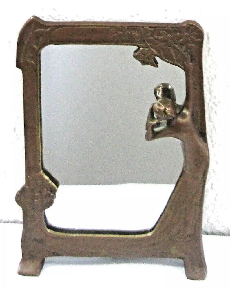 May include: A bronze mirror frame with an Art Nouveau design featuring a woman holding a flower. The frame has a decorative floral pattern and stands on a base.
