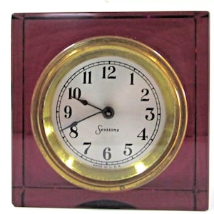 May include: A square, burgundy-colored clock with a gold-tone circular clock face. The clock face has black numerals and hands, with the word "Sessions" printed in the center. The clock is marked "MADE IN U.S.A."