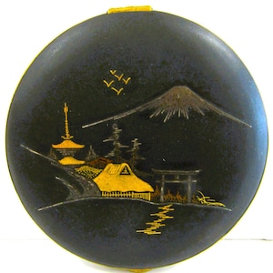 May include: A round, black compact with a gold rim and clasp. The lid features a gold and silver landscape design with a mountain, pagoda, and a thatched-roof building. The design is reminiscent of Japanese art.