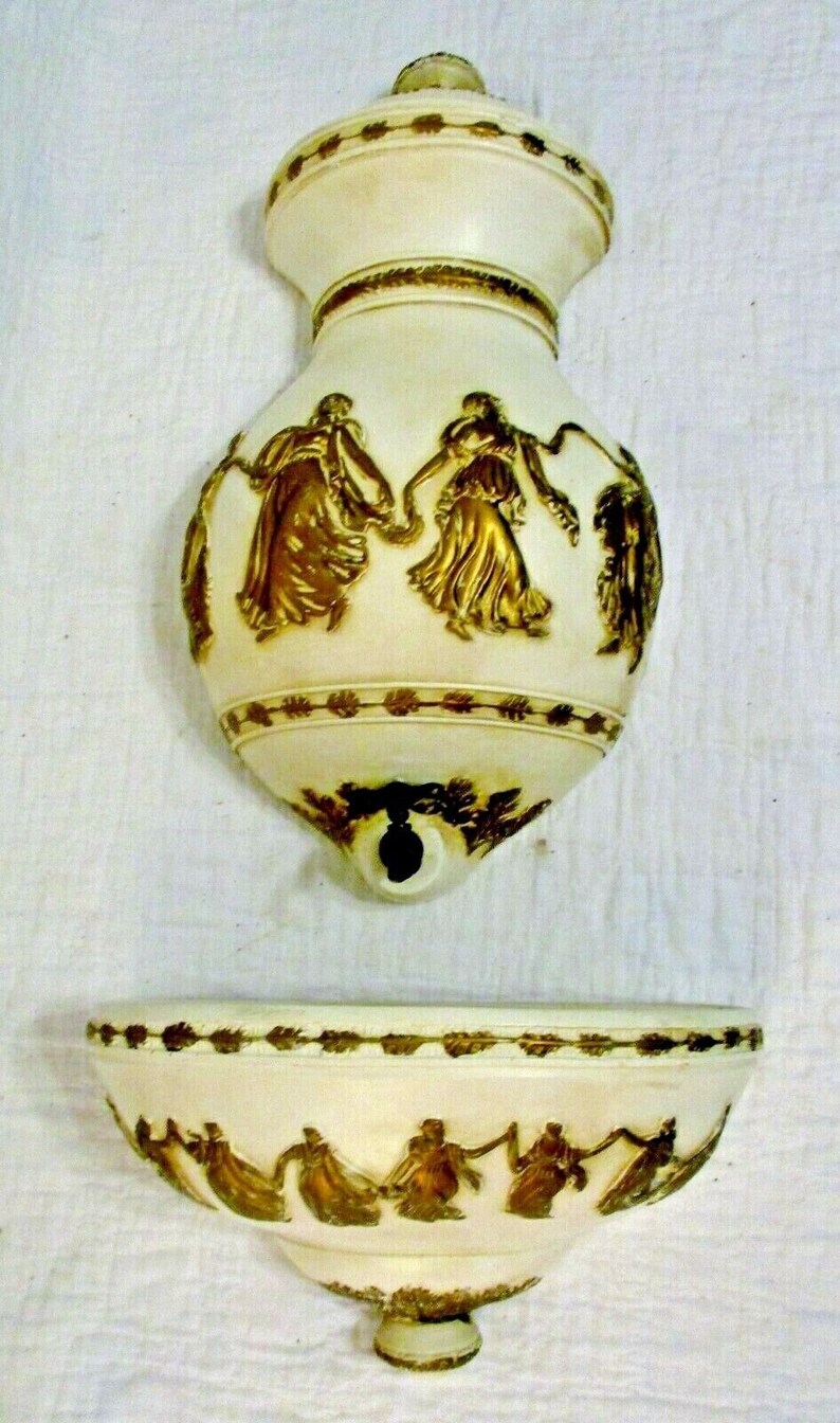 May include: A white ceramic wall fountain with gold accents. The fountain features a decorative top with a spout and a lower basin with a design of dancing figures.
