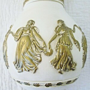 May include: White ceramic vase with gold embossed figures of women in flowing robes. The vase has a gold leaf trim around the bottom.