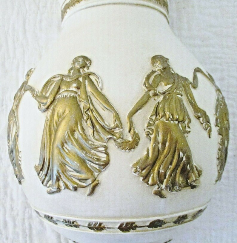 May include: White ceramic vase with gold embossed figures of women in flowing robes. The vase has a gold leaf trim around the bottom.