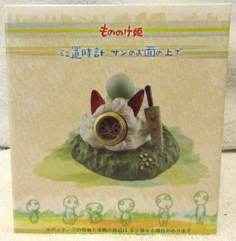 May include: A white and red figurine of the forest spirit from Princess Mononoke, sitting on a green base with a gold clock face. The figurine is a clock and the text on the box reads "もののけ姫 ミニ置時計 サンのお面の上で" which translates to "Princess Mononoke Mini Clock Sun's Face On Top".