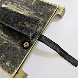 May include: The back of a gold-colored picture frame with a black felt backing and a black ribbon attached. The frame has a screw in the back.