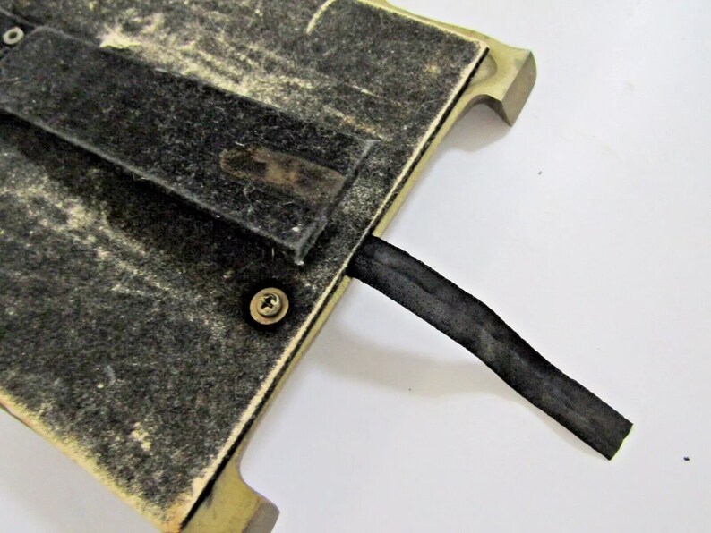 May include: The back of a gold-colored picture frame with a black felt backing and a black ribbon attached. The frame has a screw in the back.