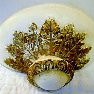 May include: A white and gold decorative ceiling medallion with a leaf design. The medallion has a raised, ornate center piece.