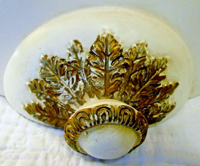 May include: A white and gold decorative ceiling medallion with a leaf design. The medallion has a raised, ornate center piece.