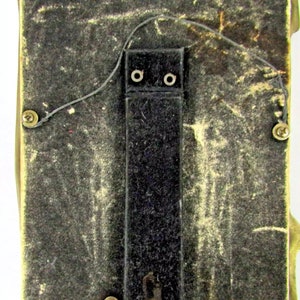 May include: The back of a brass picture frame with a black felt backing and a black hanging wire. The frame has four small brass feet.