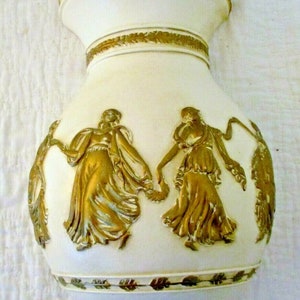 May include: White ceramic wall mounted water dispenser with gold accents and a design of two women dancing. The dispenser has a spout on the bottom.
