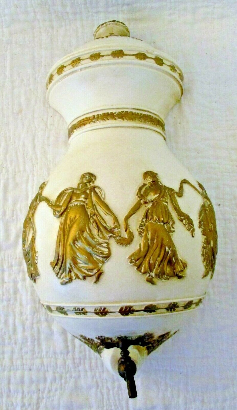 May include: White ceramic wall mounted water dispenser with gold accents and a design of two women dancing. The dispenser has a spout on the bottom.