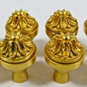 May include: Eight gold-colored cabinet knobs with a floral design. Each knob features a detailed, petal-like pattern on the top and a rounded base. The knobs are arranged in two rows, showcasing their decorative and functional design.