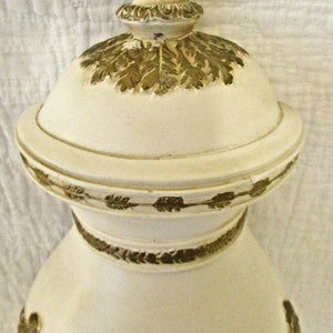 May include: White ceramic vase with gold leaf accents. The vase has a round body and a lid with a decorative knob. The lid is decorated with a gold leaf design.