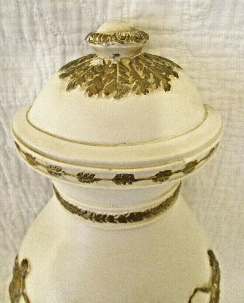 May include: White ceramic vase with gold leaf accents. The vase has a round body and a lid with a decorative knob. The lid is decorated with a gold leaf design.