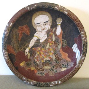 May include: A round, antique ceramic plate featuring a detailed illustration of a seated figure with a halo. The figure is dressed in patterned robes, with a dark background and decorative border. The plate has a brown and gold color scheme.