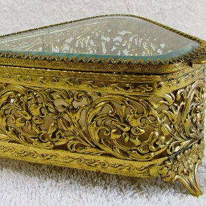 May include: A gold-tone, triangular-shaped jewelry box with a glass top and intricate floral design. The box has a hinged lid and sits on four feet.