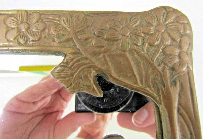 May include: A brass mirror frame with an ornate floral design. The frame is a light gold color and has a raised design of flowers and leaves.