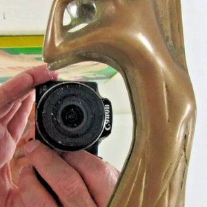 May include: A gold-toned metal mirror frame with an ornate design featuring a stylized tree and a woman's figure. The mirror is reflecting a hand holding a camera.