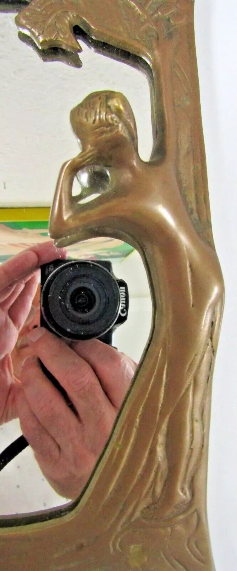 May include: A gold-toned metal mirror frame with an ornate design featuring a stylized tree and a woman's figure. The mirror is reflecting a hand holding a camera.