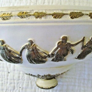 May include: White ceramic bowl with a gold trim and a raised design of figures in flowing robes. The bowl has a rounded base.