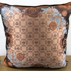 May include: Square decorative pillow with a brown and tan geometric pattern. The pillow features a repeating pattern of a Greek key design and a Medusa head. The edges are trimmed with a dark brown rope.