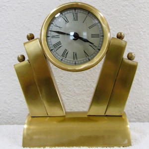 May include: A gold-toned desk clock with Roman numeral markings on the face. The clock is on a decorative stand with a geometric design.