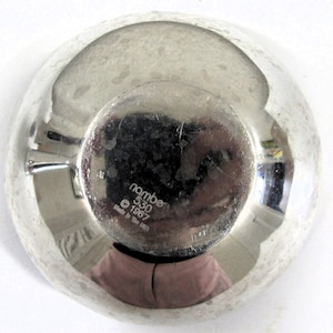 Puede incluir: Un cuenco de plata con una superficie reflectante. La parte inferior del cuenco tiene el texto "nambe 530 &copy; 1967 Made in the USA".