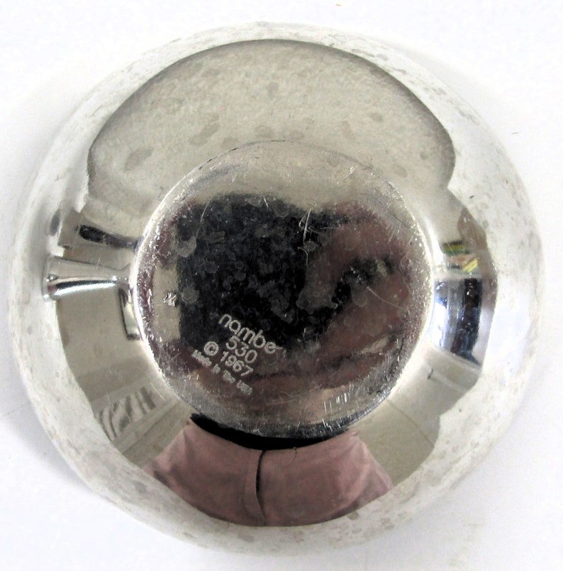 Puede incluir: Un cuenco de plata con una superficie reflectante. La parte inferior del cuenco tiene el texto "nambe 530 &copy; 1967 Made in the USA".