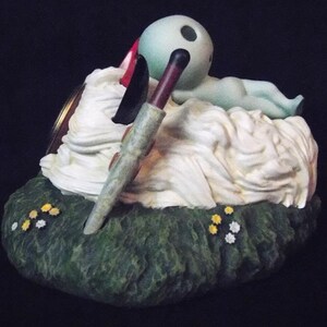 May include: A green and white figurine of a creature lying on a bed of white clouds. The creature has a large head with two black eyes and a long, thin body. The figurine is sitting on a green base with yellow flowers.