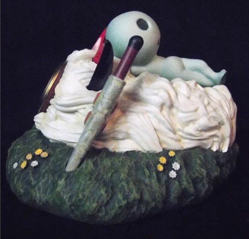 May include: A green and white figurine of a creature lying on a bed of white clouds. The creature has a large head with two black eyes and a long, thin body. The figurine is sitting on a green base with yellow flowers.