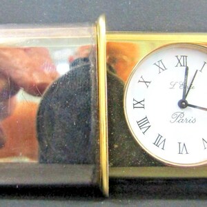 May include: A rectangular travel clock with a gold-colored frame and a white clock face with black Roman numerals. The clock face reads "L'Etoile Paris". The clock is encased in a clear, rectangular case.