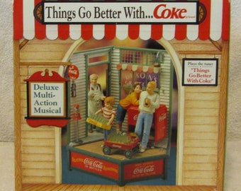 May include: A Coca-Cola themed musical toy set in a miniature town setting. The toy features a red wagon, a Coca-Cola vending machine, and a small store with a sign that reads "Dixon's Grocery". The toy plays the tune "Things Go Better With Coke".