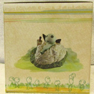 May include: A small, gray, figurine of a creature with large eyes and a long, thin body, sitting on a white, fluffy cloud. The figurine is surrounded by a green, grassy area. The background is a light brown with a pattern of yellow and green stripes.