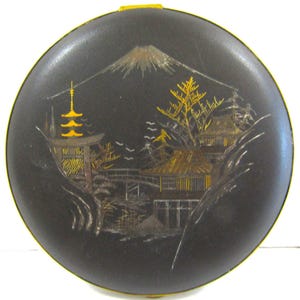 May include: A round, black compact with a gold-colored rim. The lid features a detailed landscape scene with a mountain, pagoda, and buildings, all in gold and silver tones. The compact is likely vintage.