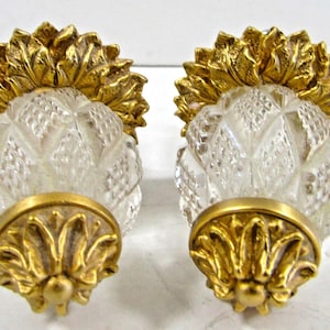 May include: A pair of decorative cabinet knobs. Each knob has a clear, diamond-cut glass body, topped with a gold-coloured, stylised floral design. The base of each knob has a matching gold-coloured floral accent.