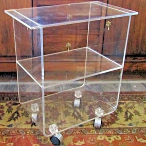May include: A clear acrylic rolling cart with two shelves and small gold-colored decorative accents. The cart has a rectangular top and a curved bottom, with clear cube-shaped feet and small wheels for mobility. The cart is designed for storage and display.