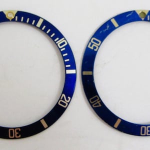 May include: Two blue watch bezels with white and gold markings. The bezels have numbers from 10 to 50, with a gold triangle at the top. The bezels are circular and appear to be watch parts.
