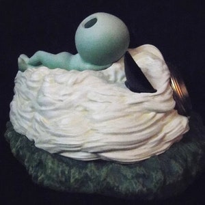 May include: A ceramic figurine of a green alien laying on a white cloud with a black and gold lid on top.