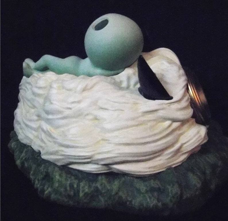 May include: A ceramic figurine of a green alien laying on a white cloud with a black and gold lid on top.