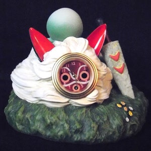 May include: A white ceramic clock shaped like a No Face mask from the movie Spirited Away. The clock has a red face with black hands and a gold rim. The mask is sitting on a green base with white flowers.
