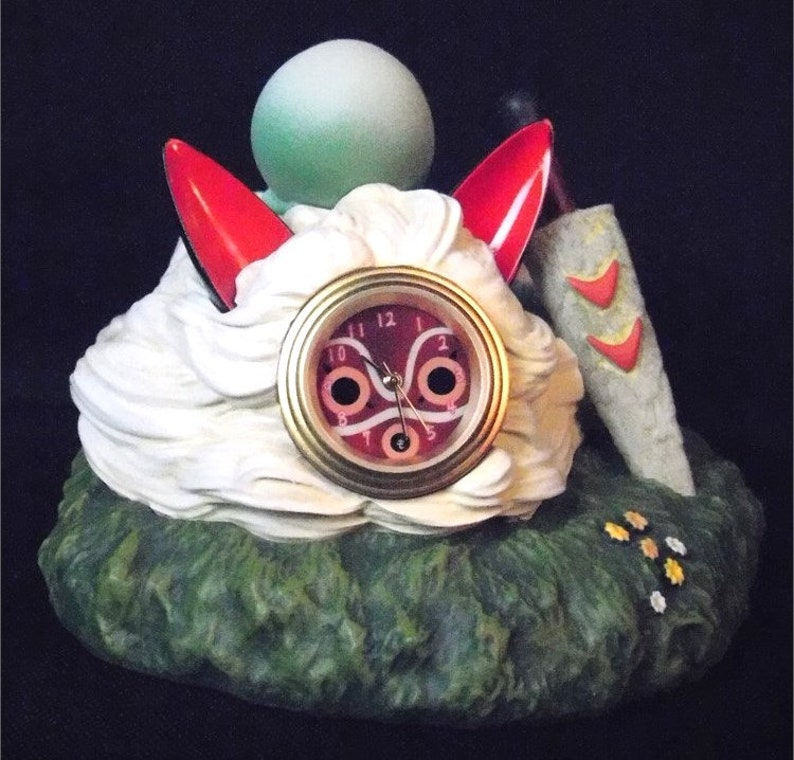 May include: A white ceramic clock shaped like a No Face mask from the movie Spirited Away. The clock has a red face with black hands and a gold rim. The mask is sitting on a green base with white flowers.