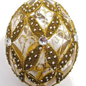 May include: A clear glass egg-shaped ornament featuring a gold glitter lattice pattern with textured gold bead accents and small silver glitter dots. The design includes clear faceted circular crystals at the intersections and a gold cap.