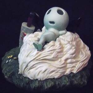 May include: A small, light blue figurine of a character from the movie 'My Neighbor Totoro' is sitting on a white, fluffy cloud. The figurine is wearing a white dress and has a big, round head with large eyes. The cloud is sitting on a green base.