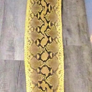 May include: A long, tanned snakeskin with a yellow and brown pattern. The skin is laid out on a gray wooden surface, showcasing its natural markings and textures. The snake skin is a natural product.